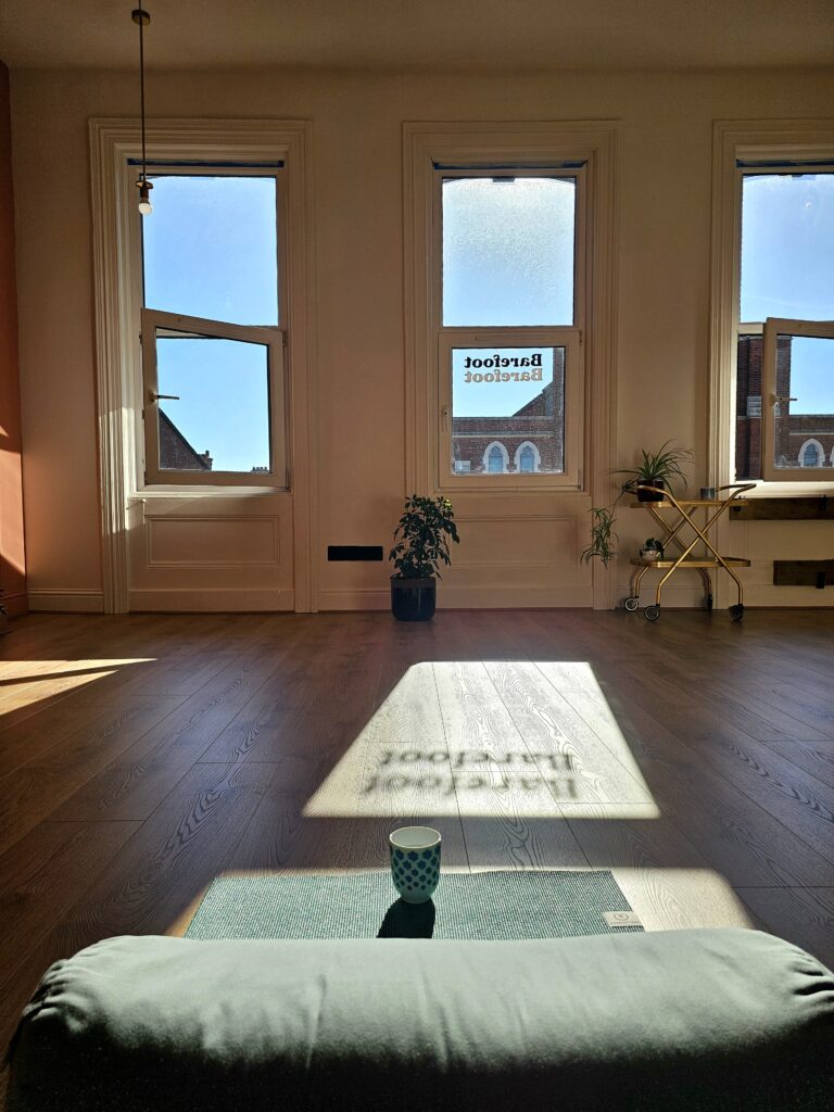 A water cup, yoga mat and bolster sit on the studio floor with the dawn sunlight casting the shadow of the studio name on the floor: 'Barefoot'